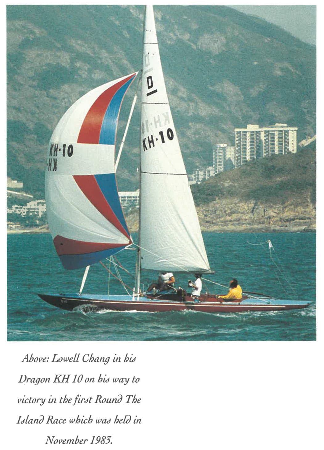 A sailboat with red, white and blue sails, with a hilly landscape and tall buildings behind.