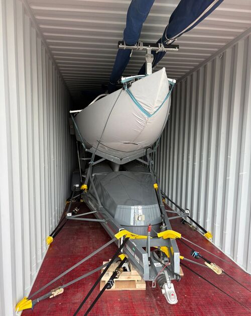 Two Dragon hulls stacked and strapped inside a shipping container, masts lashed to the roof and the trailer hitch resting on timber blocks