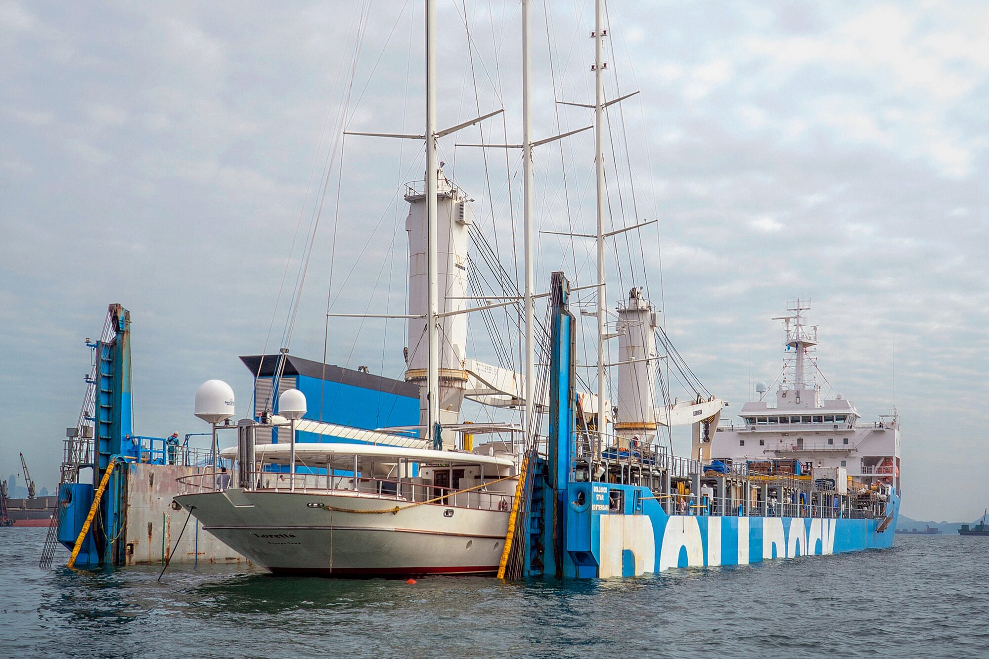 The three-masted yacht Loretta loaded aboard the heavy-lift ship Rolldock Star in harbour, with port cranes and a green island behind
