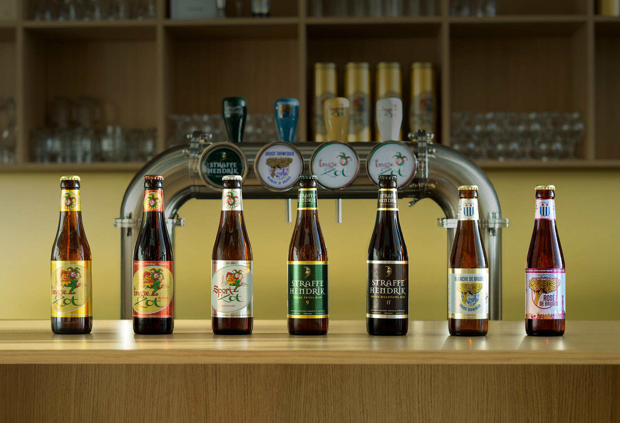 Seven De Halve Maan bottles lined up along a bar in front of the taps, among them Brugse Zot, Sport Zot, Straffe Hendrik and Blanche de Bruges