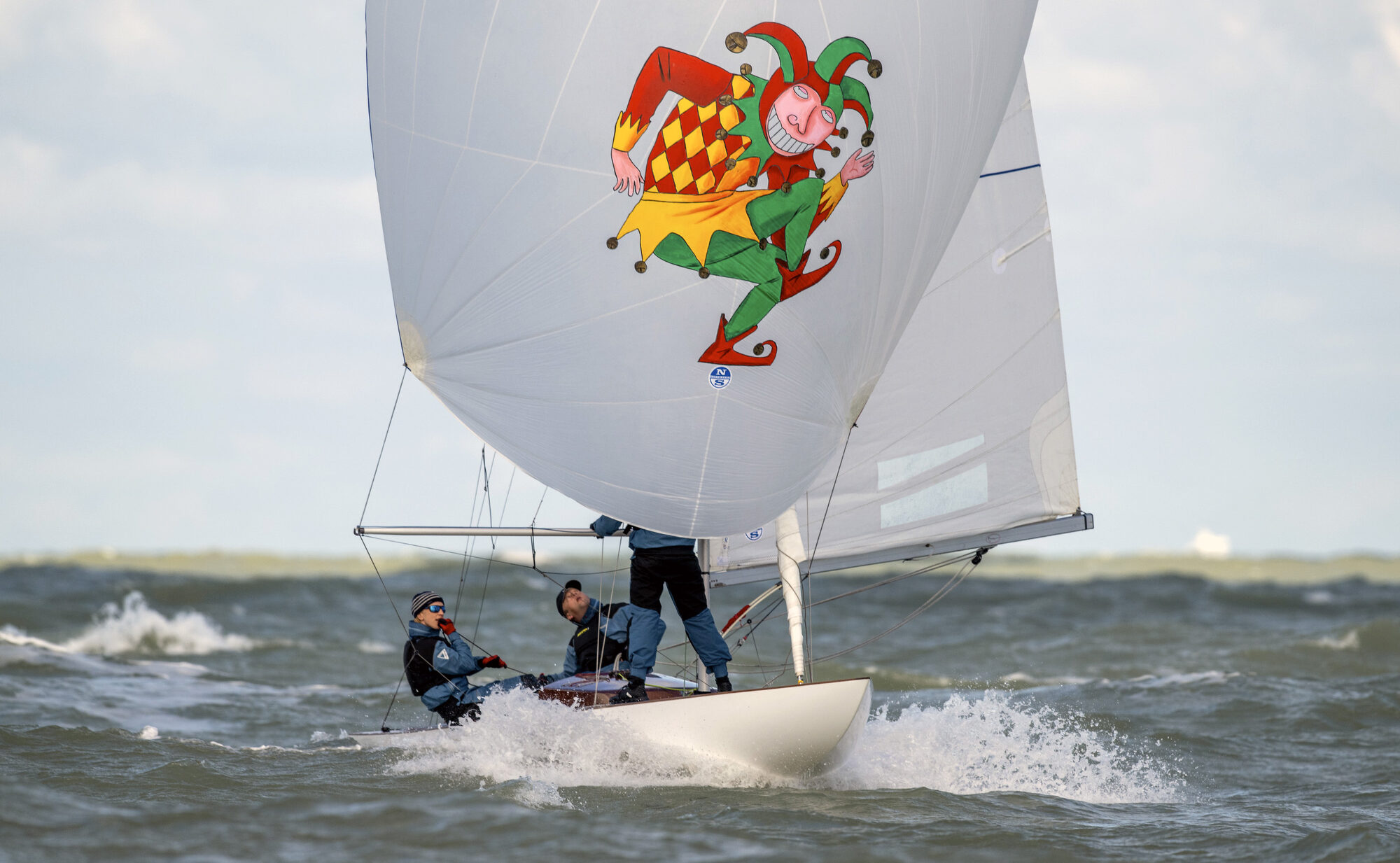 A Dragon reaching hard under a white spinnaker carrying the Brugse Zot jester, its three crew on the rail as the bow throws spray