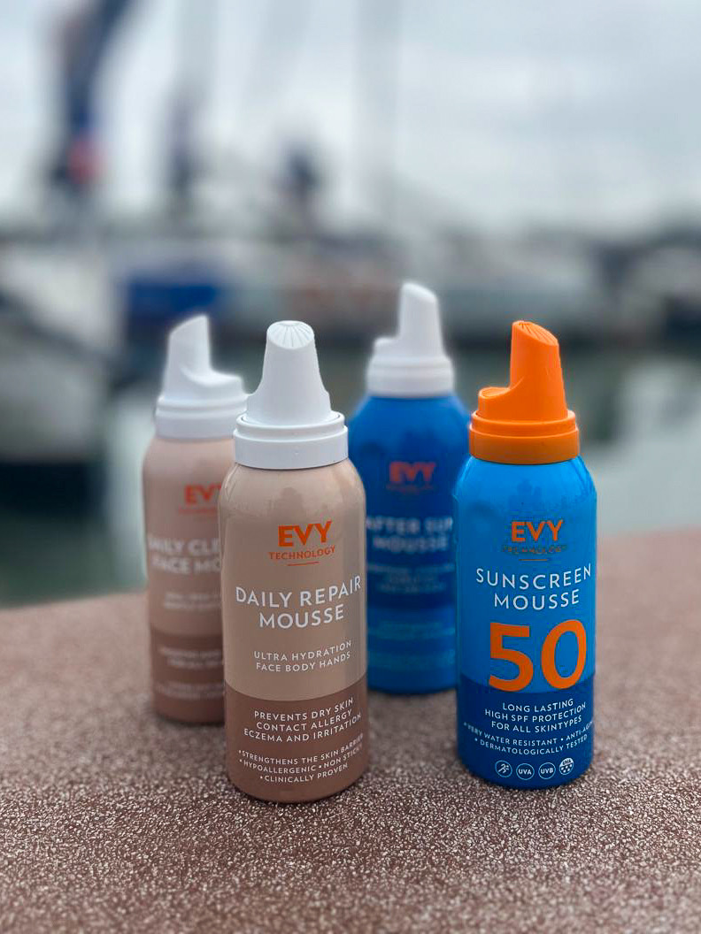 Four EVY Technology mousse cans standing on a harbour wall, among them Sunscreen Mousse 50 and Daily Repair Mousse, with masts out of focus behind