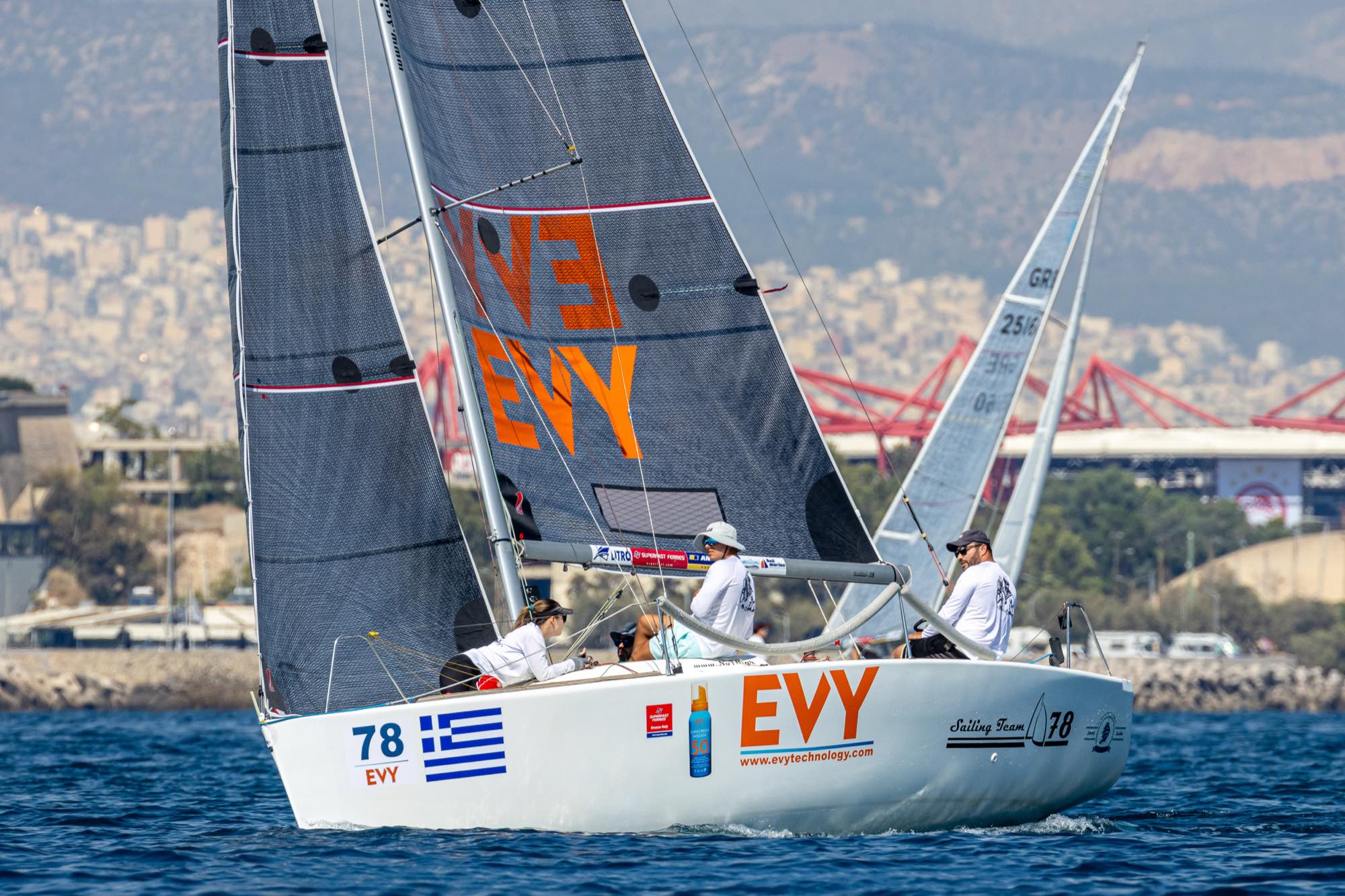 A keelboat carrying EVY Technology's orange wordmark on its mainsail and topsides, four crew aboard, racing off Piraeus with the city behind