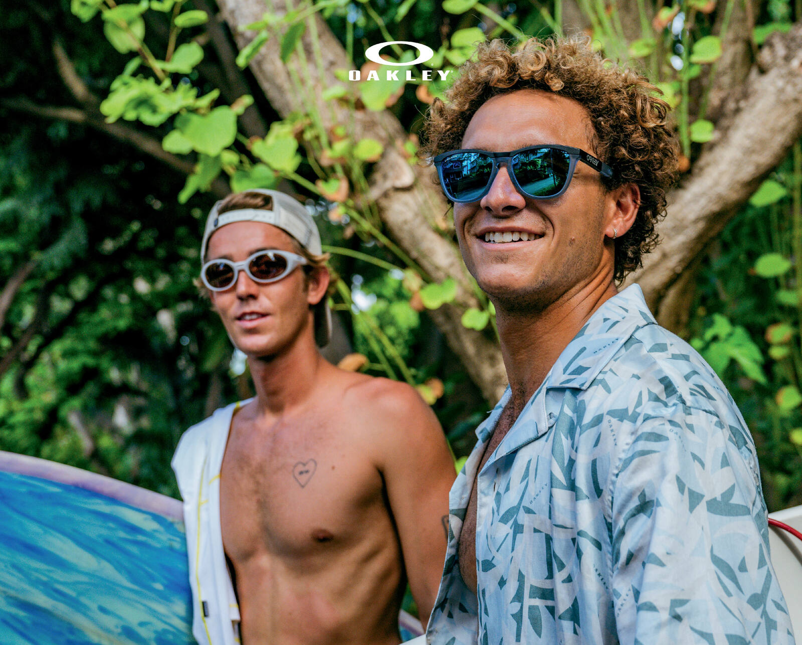 Two surfers standing under trees with their boards, one wearing dark Oakley sunglasses with mirrored lenses, the other white-framed sunglasses