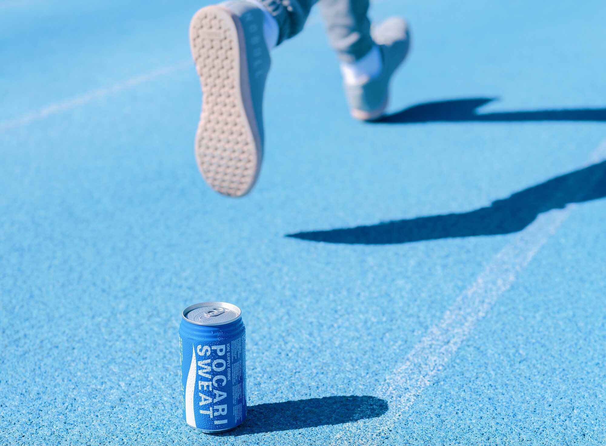 A can of Pocari Sweat standing on a blue running track, a runner’s shoe in the air above it