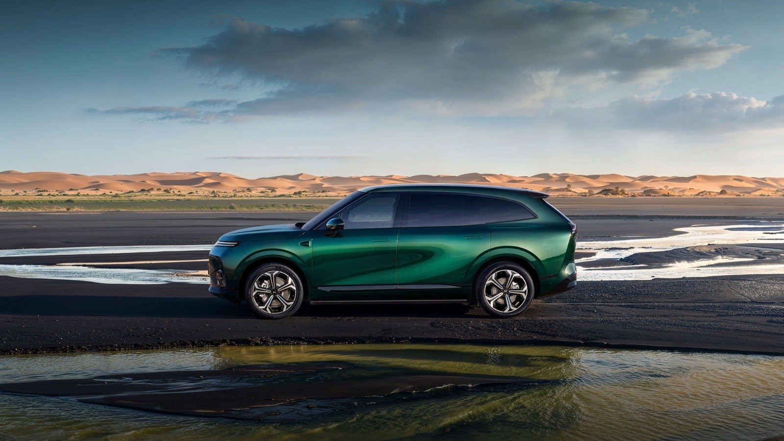 The XPENG G9L electric SUV in dark green, parked in profile on a wet salt flat with desert dunes behind it