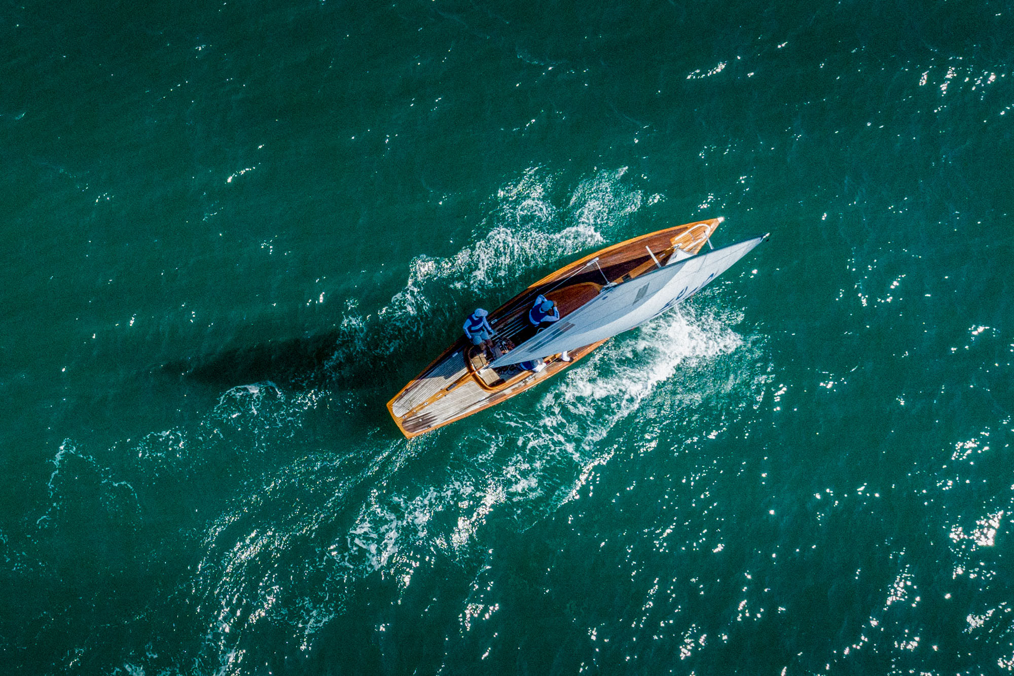 A varnished wooden Dragon seen from directly above, two crew aboard, cutting a white wake across deep green water.
