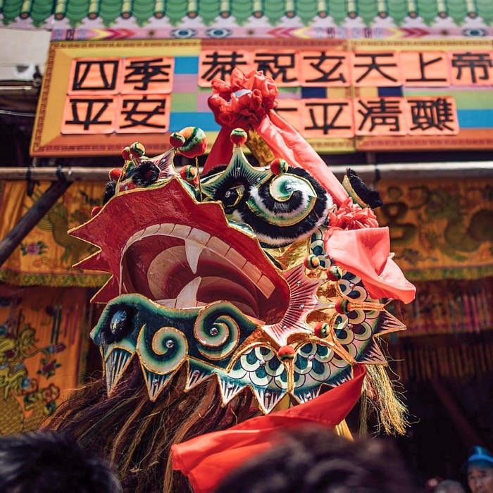 A dragon dance head in red, gold and green, raised in front of a decorated temple at a Hong Kong festival.