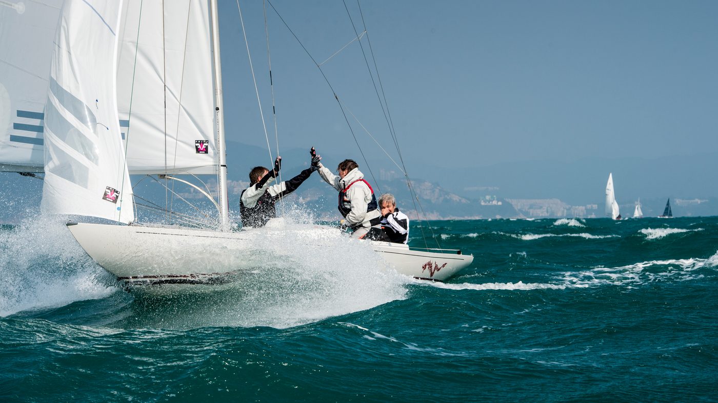 Three sailors high-five aboard a Dragon keelboat as it drives through spray, other boats and a hazy coastline behind.