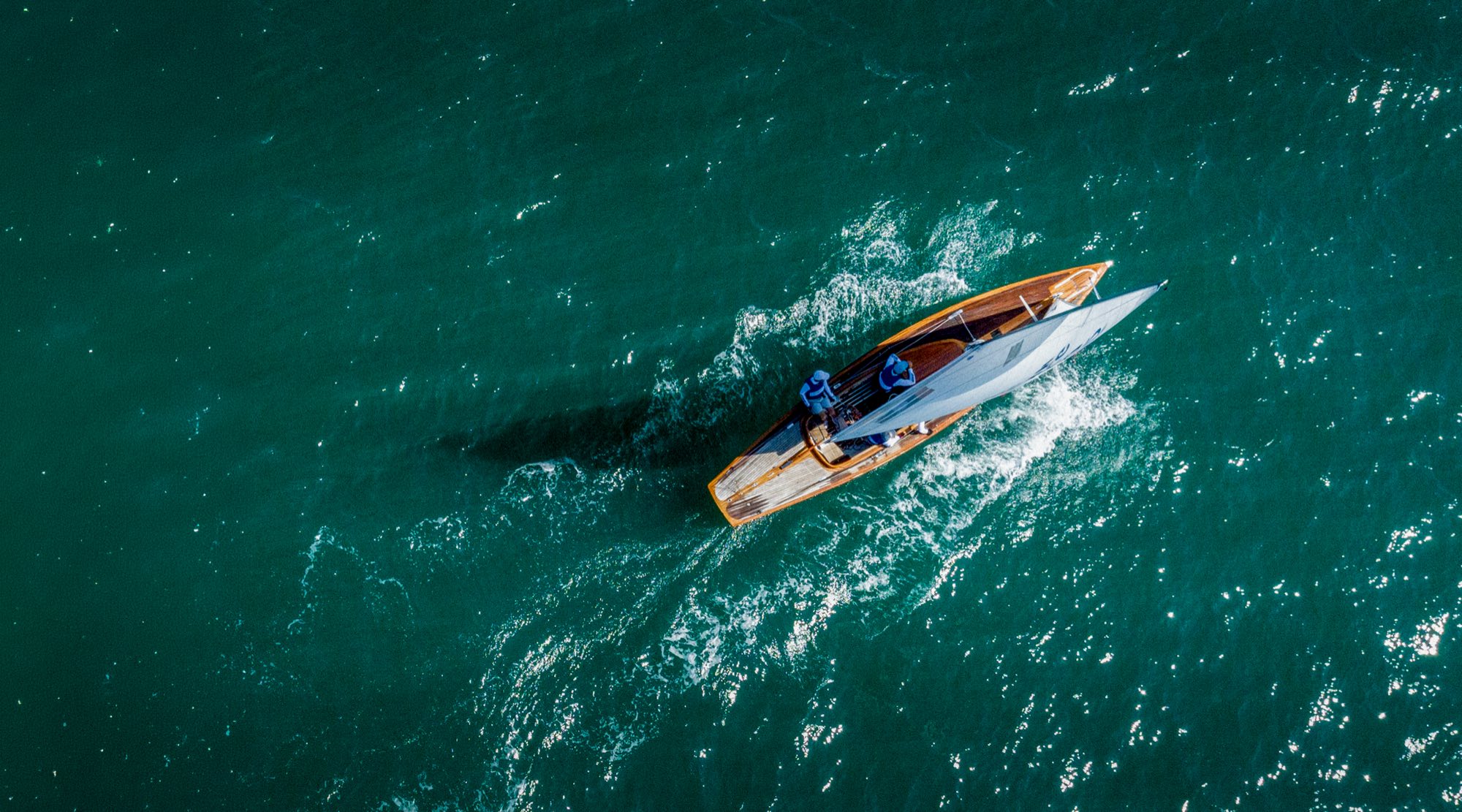 A Dragon keelboat seen from directly above, its varnished wooden hull and white sail cutting a wake across open green water off Hong Kong