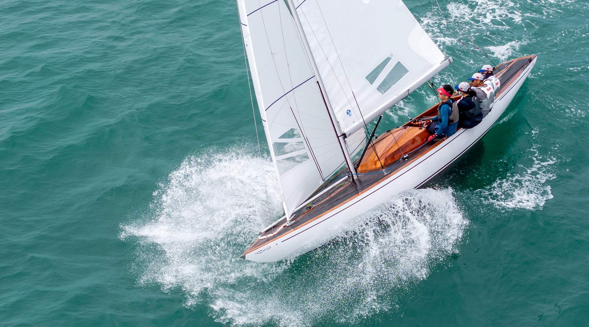 A Dragon keelboat with a varnished teak deck powering upwind, its crew hiking out, seen from above off Hong Kong