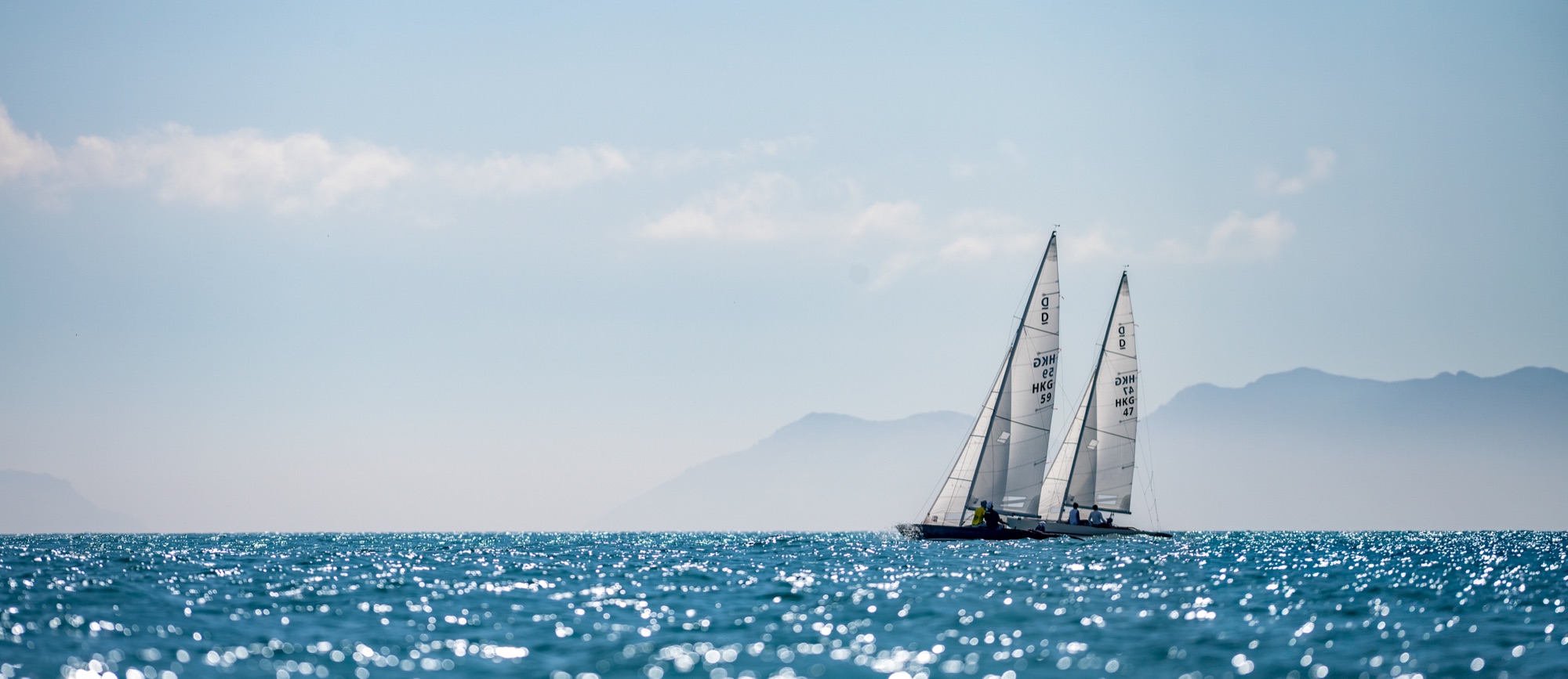 Two Hong Kong Dragons sailing close together on sunlit open water, the hills of the outlying islands hazy behind them.