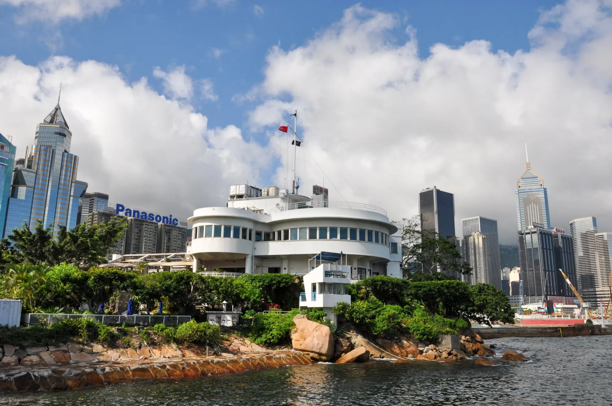 A white waterfront clubhouse with green trees and a city skyline behind.